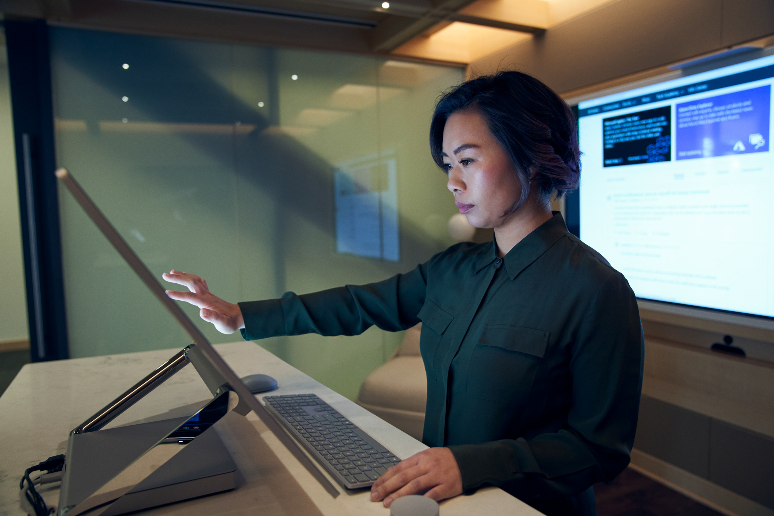 Side profile of a woman wearing a dark shirt in a dim office scrolling or working on a Microsoft Surface Studio. Keywords: touch screen, desktop, cloud security, threat protection, secure score, monitoring, Microsoft Security collection