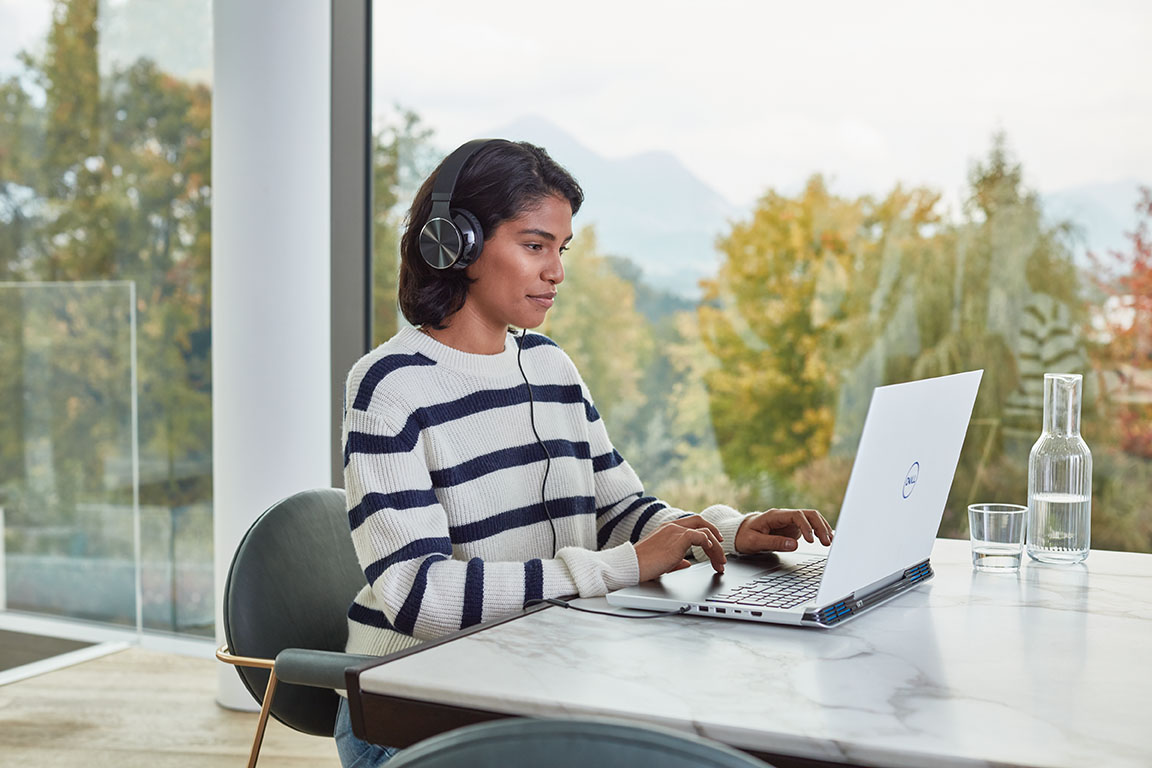 a woman sitting at a table with headphones on looking at a white laptop.