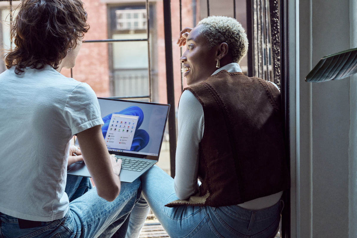 Two Gen Z friends sitting on fire escape balcony with backs to camera laptop open with Windows 11 start screen and start menu.