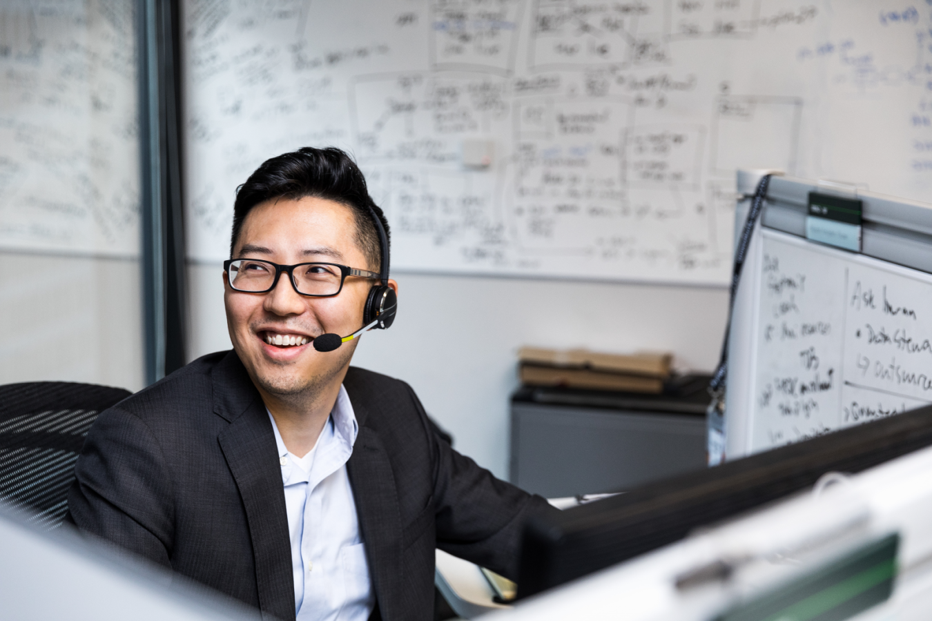 Male worker sitting at cubicle desk in financial office, wearing glasses and phone headset. He is smiling and looking up to his right. A large whiteboard covered in black written text is on wall behind him. A smaller whiteboard also partially visible on his desk‘s cubicle wall.