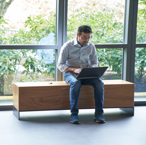 Man sitting on an indoor bench in front of windows working on a laptop with a security key fob or USB drive next to him on the bench.