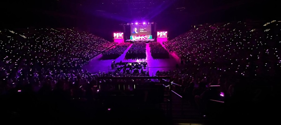 A wide shot of an arena full of attendees at a the 2023 Power Platform Community Conference; the room is dark except for the large presentation screens at the stage.
