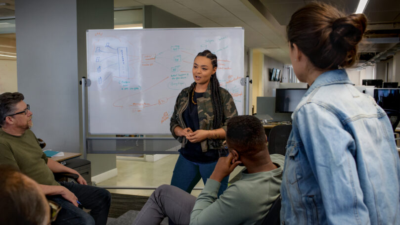Team members gathered in an open office, listening to a colleague presenting ideas on a whiteboard.