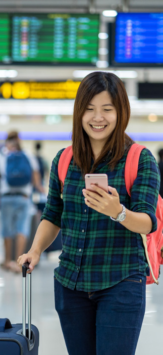 Asian woman looking at her cellphone pulling a rolling suitcase and wearing a backpack in an airport