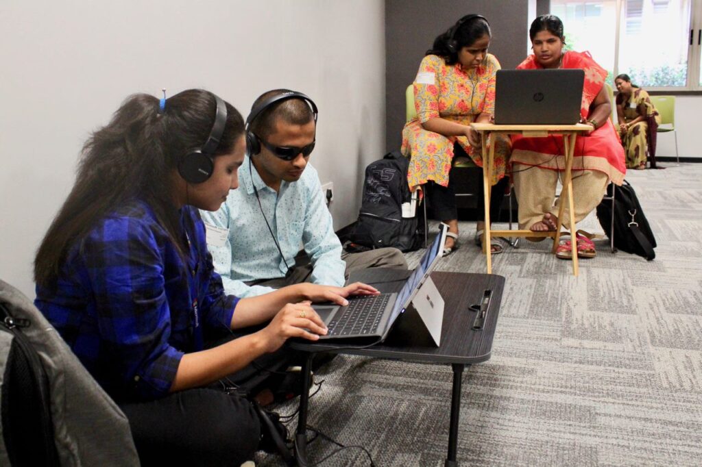 A woman and man both wearing headphones connected to a laptop on a small table in front of them are seated on a carpeted floor. The woman has her hands on the keyboard. Behind them, two women, one wearing headphones, are seated at a folding table, their attention on a laptop in front of them. A backpack and messenger bag rest on the floor beside them. 