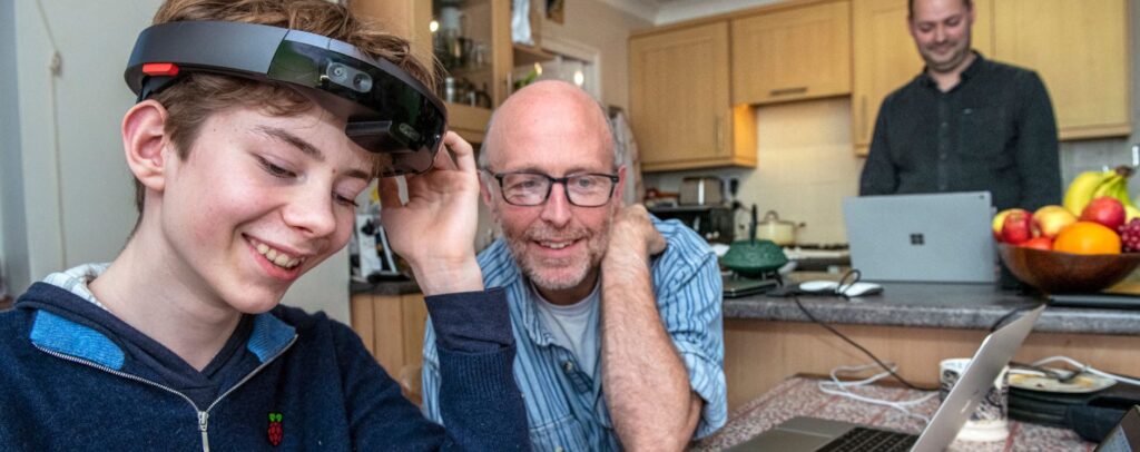 A smiling young boy who is blind and wearing a head-mounted augmented reality device with a semicircular LED interface is seated at a kitchen table. A man, also smiling, is seated next to him, a laptop in front of him. In the background, another man is standing behind a kitchen island, working on a laptop. 
