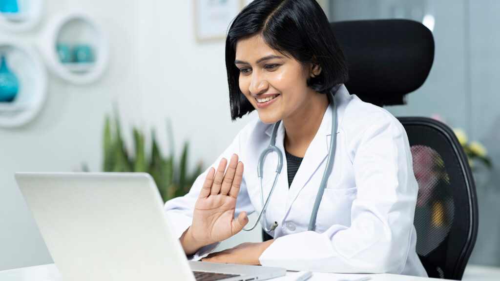 a female doctor sitting at a table using a laptop computer for a teleconference