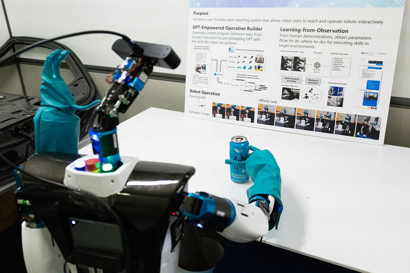 Photo of a robotic arm grasping an aluminum can on a desk; there is a presentation poster standing at the back edge of the desk
