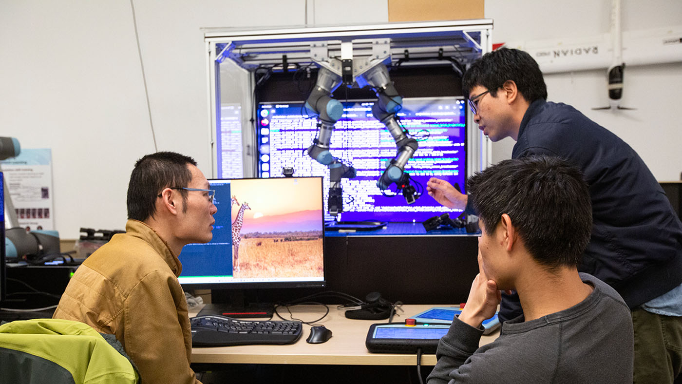 photo of three researchers collaborating on a bimanual coordination in front of two robotic arms suspended from a frame in front of a screen of code