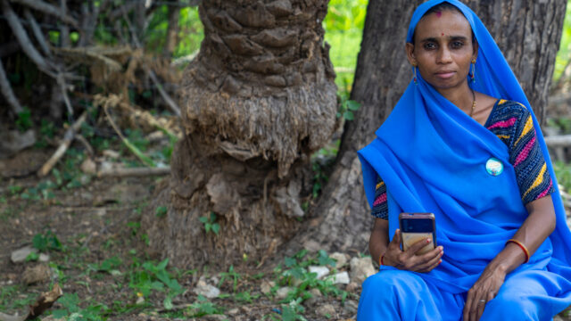 Lady in blue sari, holding a smartphone, looking at the camera. Seated outside in front of a couple of trees.