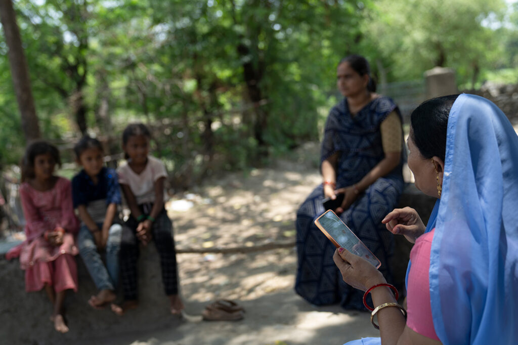 A woman in a blue sari is holding a smartphone and is sitting across from three children and a woman in a maroon sari.