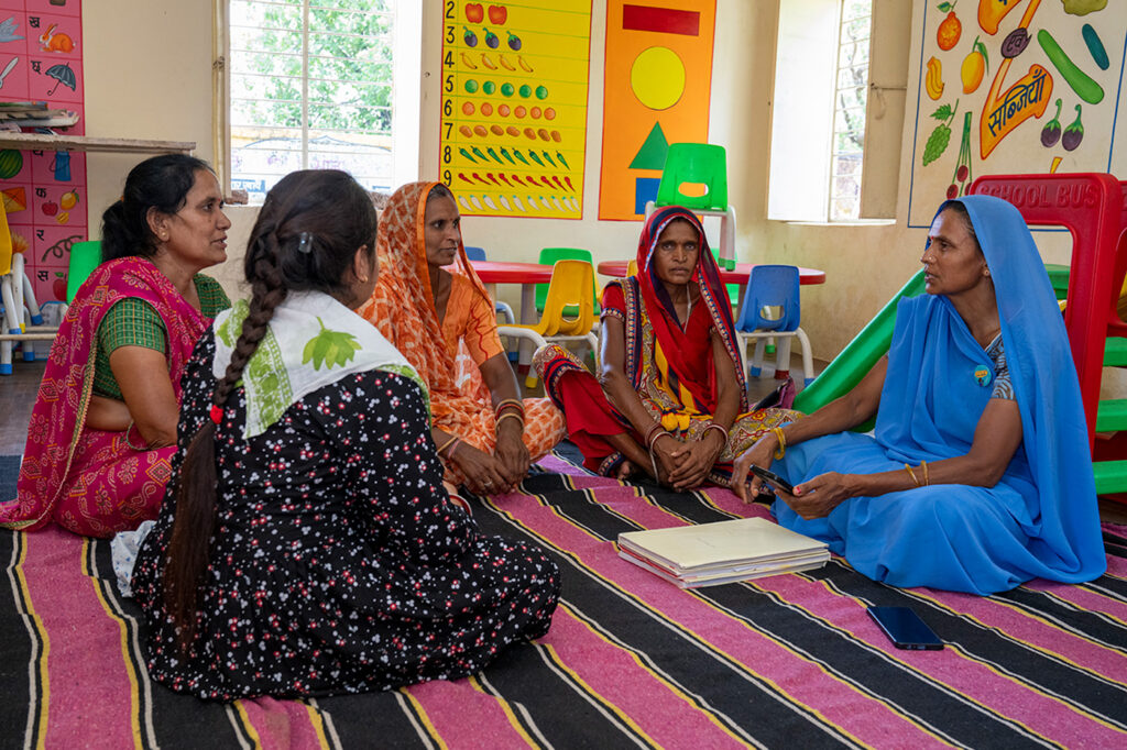 Five ladies in colorful saris seated on a rug at a classroom talking. The lady on the far right, wearing a blue sari, is holding a smartphone, and has a stack of papers at in front of her.