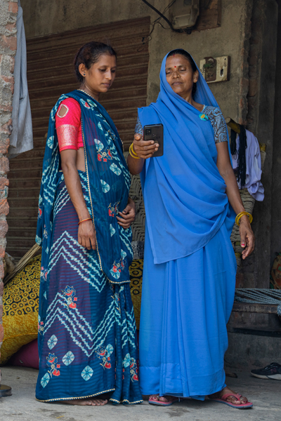 One lady in blue sari talking to ASHA standing outside of a brick house, one of them holding a smartphone.