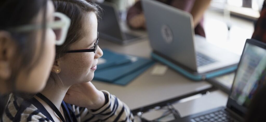 Two female middle-school students seated at a table, focused on a laptop screen. The girl in the foreground wears glasses and a striped shirt, typing with one hand while the other rests on their neck. A second girl observes from beside her. The table holds additional laptops and folders, suggesting a collaborative or educational setting. The scene conveys active engagement with technology in a learning or workshop environment.