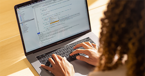 An image of a Black female developer coding on a MacBook Pro in the office, using Visual Studio.