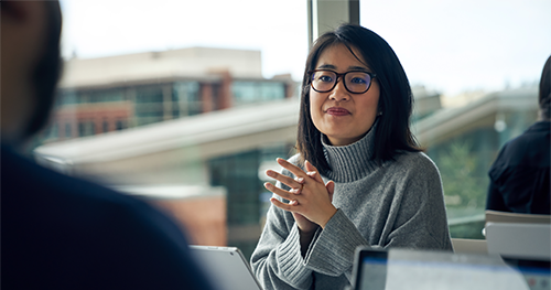 An image of a woman with glasses sitting at a community table in common area looking up from behind a Surface laptop with her hands clasped together.