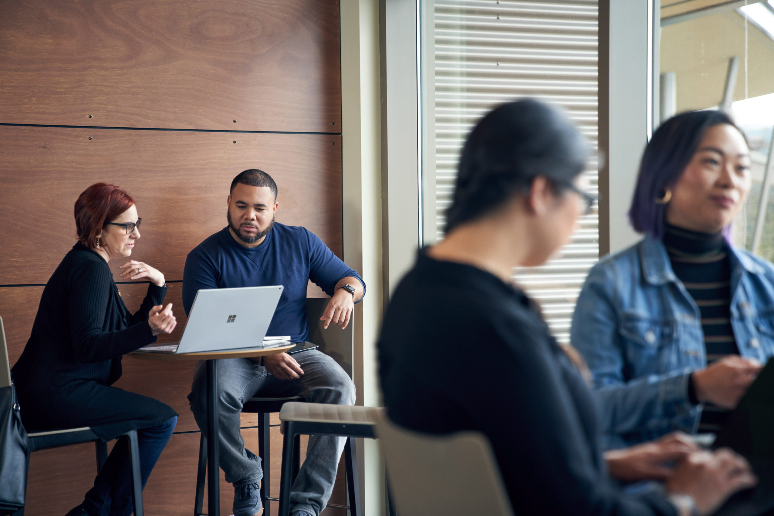 Man and woman meeting together and reviewing content on a Surface laptop at a high table with two blurred women in the foreground.