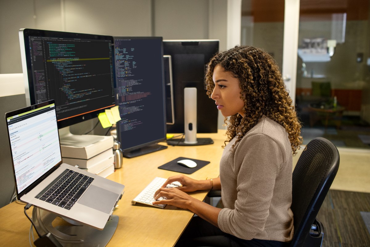 Black female developer working at enterprise office workspace. Focused work. She has customized her workspace with a multi-monitor set up.