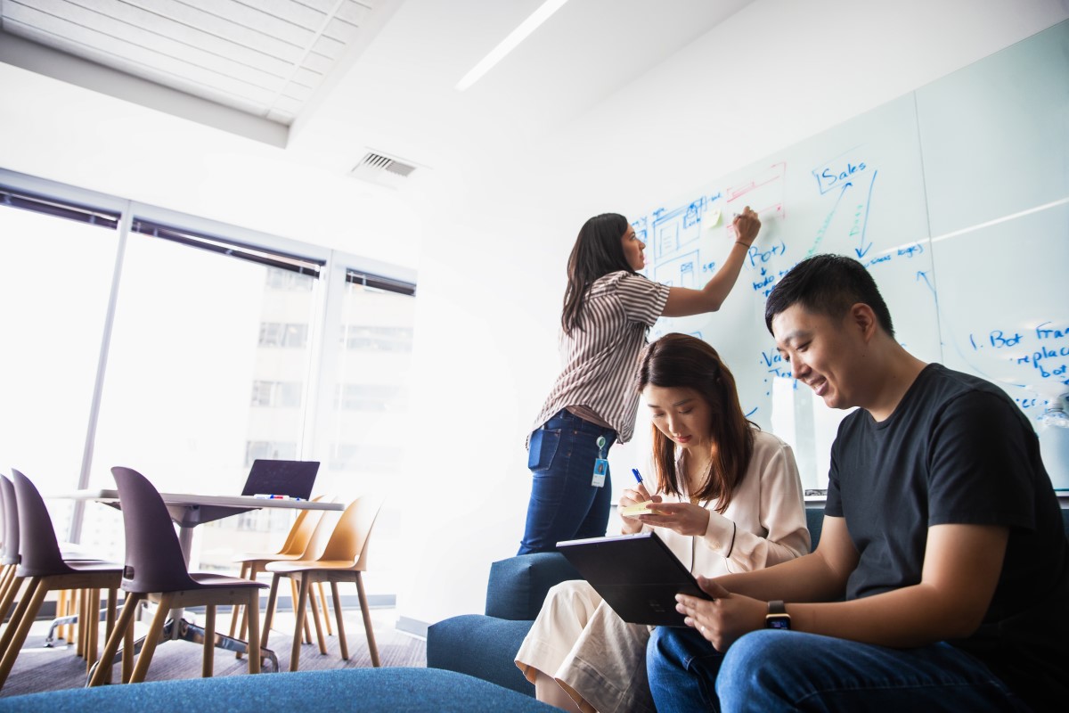 A group of coworkers collaborate on project in conference room of U.S. office. Woman writes on white board while two other coworkers work on a laptop together.
