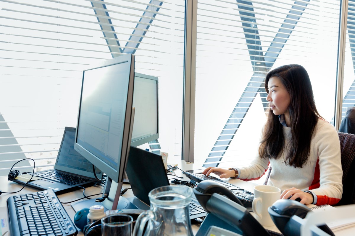 Female office worker at workstation near window, looking at desktop monitor (screen not shown). Open laptop also on desk (screen not shown).