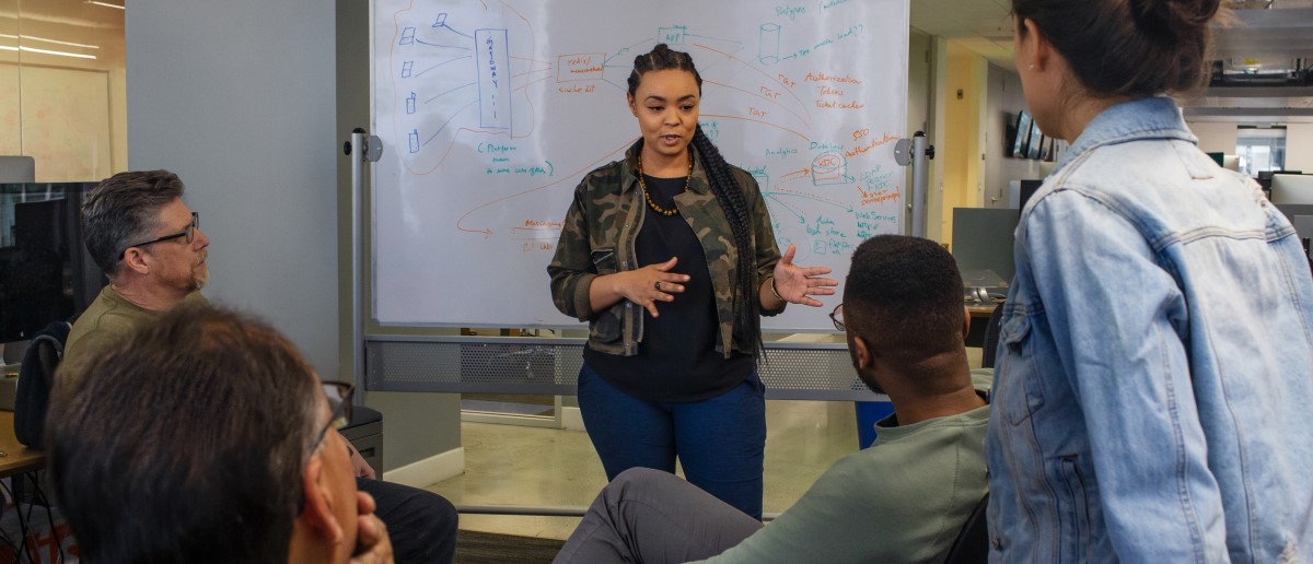 Real people, real offices. Black female developer speaking in front of a white board during team stand up meeting.