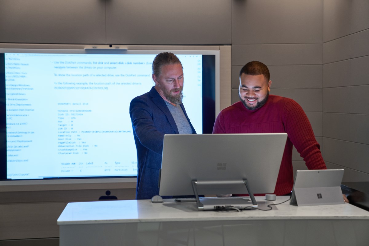 Forward facing view of two men working on a Microsoft Surface Studio with a larger blurred screen/display behind them.
