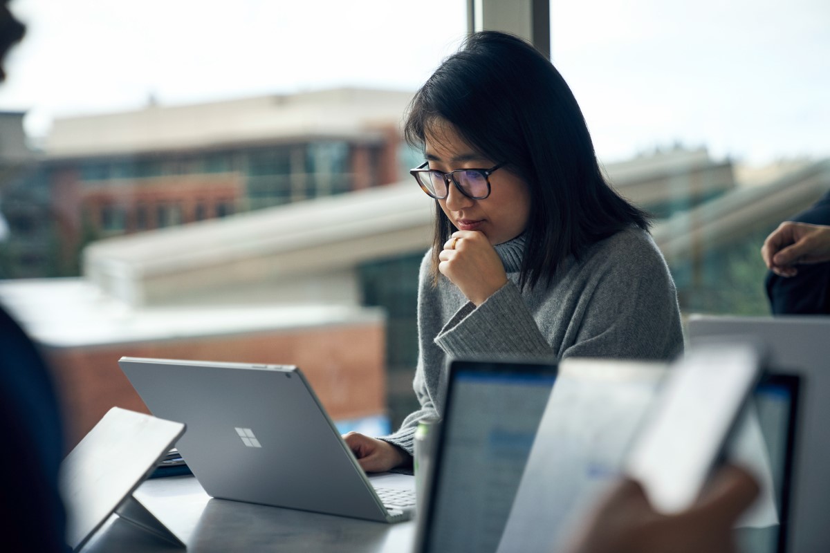 Woman with glasses sitting at a community table in common area reading on a Surface laptop. She looks as though she may be thinking, or learning.
