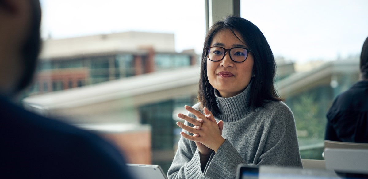 Woman with glasses sitting at a community table in common area looking up from behind a Surface laptop with her hands clasped together.