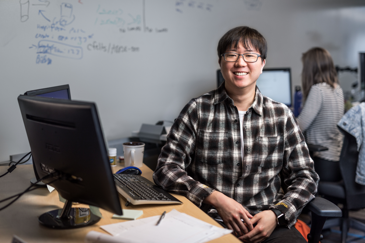 Portrait of male developer in glasses and plaid flannel shirt, smiling and facing camera. He is sitting at a desk in an office setting, with both hands on his lap. Keyboard and desktop computer visible, though screens are not shown. A female worker is shown seated at desk using computer in background (screen blurred). Whiteboard wall also shown in background with handwritten diagrams and formulas.