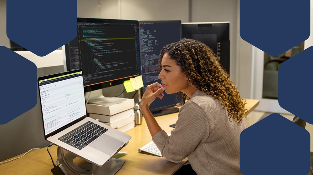 Photo of a female developer working at enterprise office workspace. Focused work. She has customized her workspace with a multi-monitor set up.