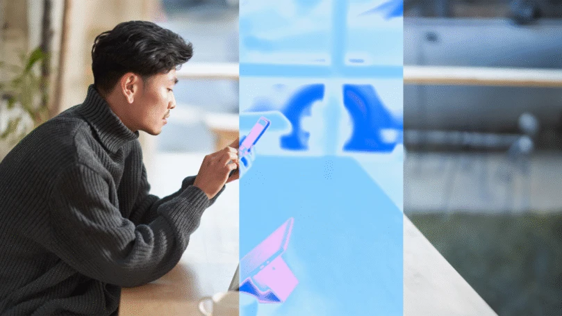 A man sitting at a laptop holding a cell phone.