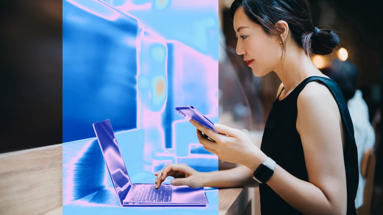 A woman holds a smartphone and types on a laptop.