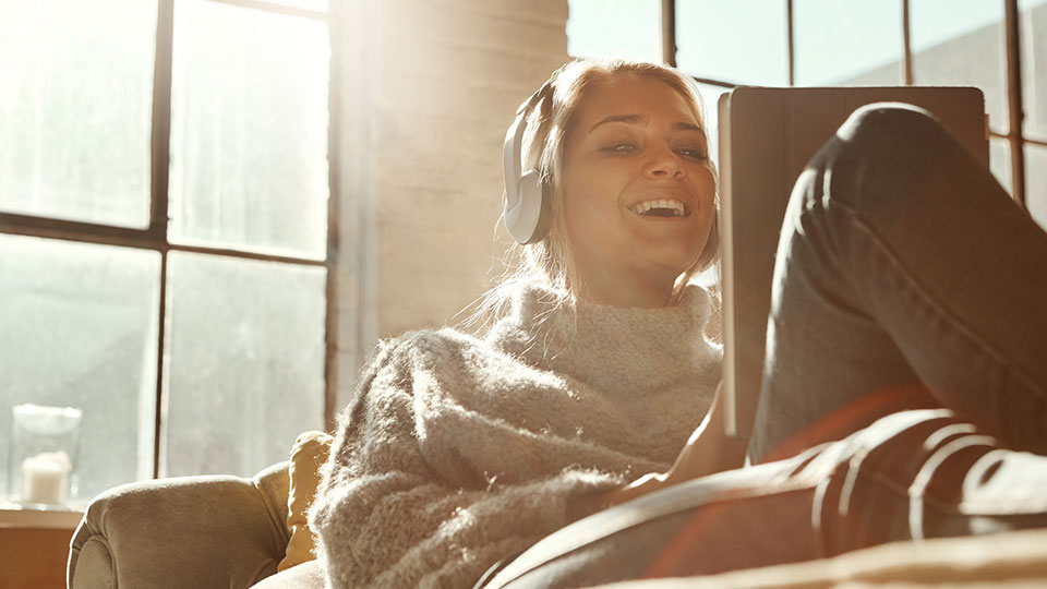 A smiling woman sits on a sunlit couch with a tablet