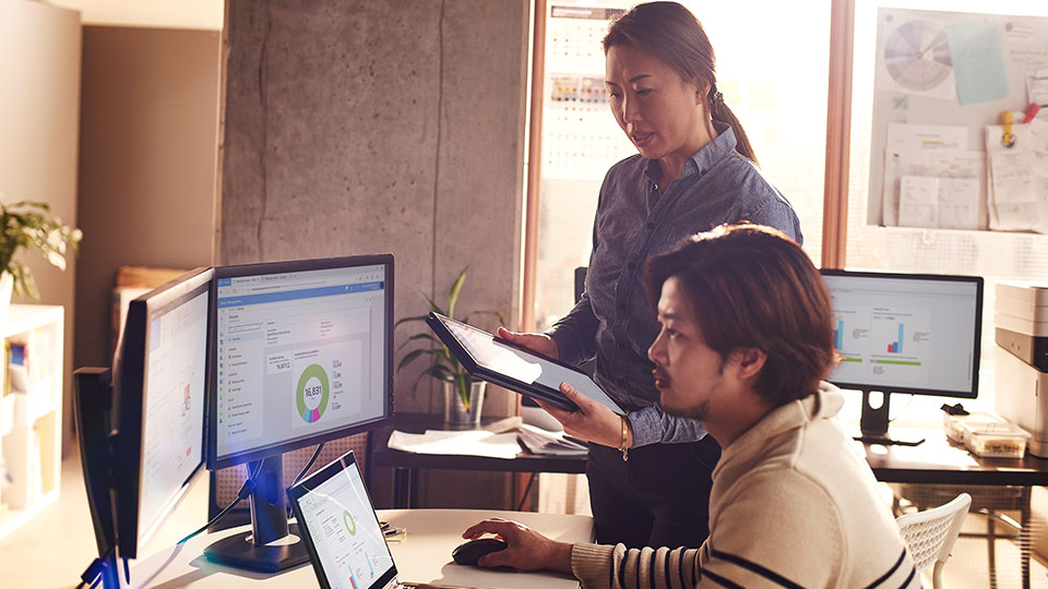 Two coworkers compare information on a tablet to information on a computer monitor