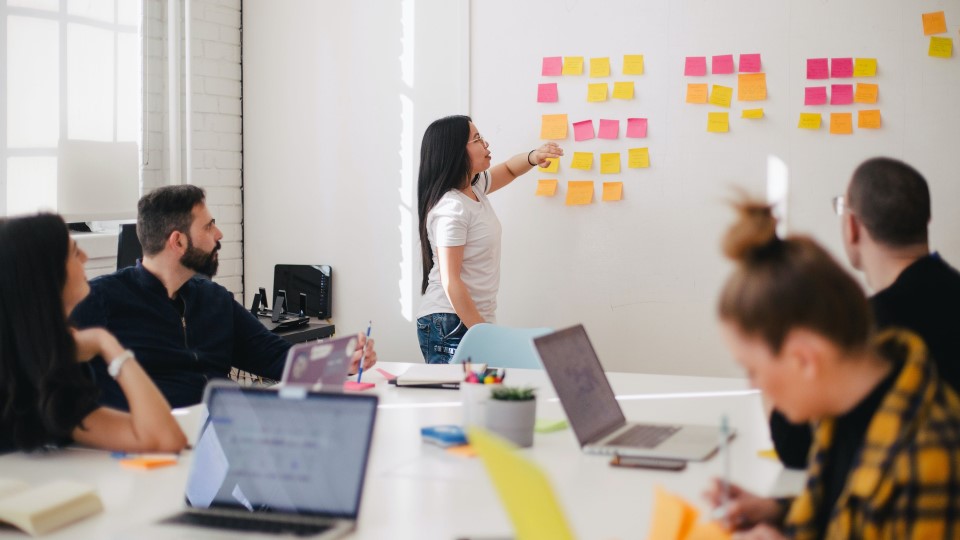 A woman points to sticky notes in a conference room as others look on.