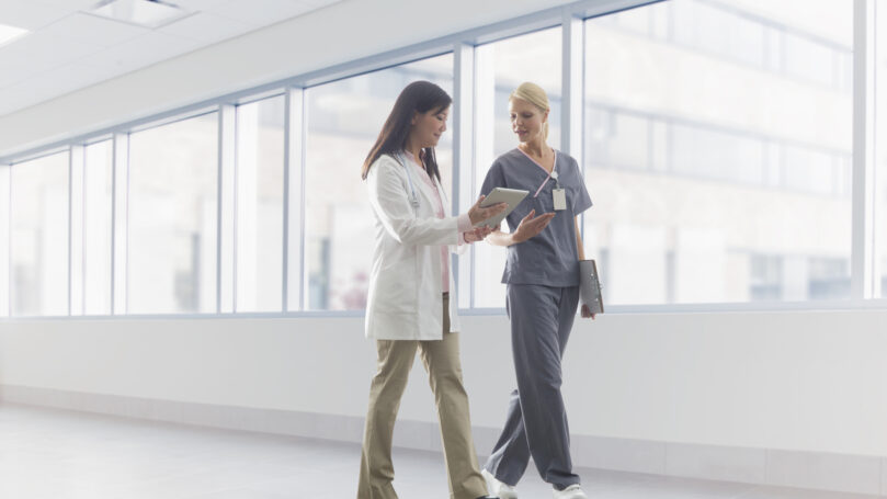 Two doctors walking down a hallway in a hospital looking at a tablet device.