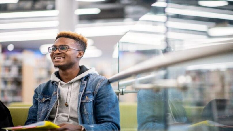 A smiling university student wearing a hoodie and jeans jacket sits in a brightly lit library.