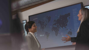 Two executives discuss security challenges in an office setting in front of a large screen showing a map of the world.
