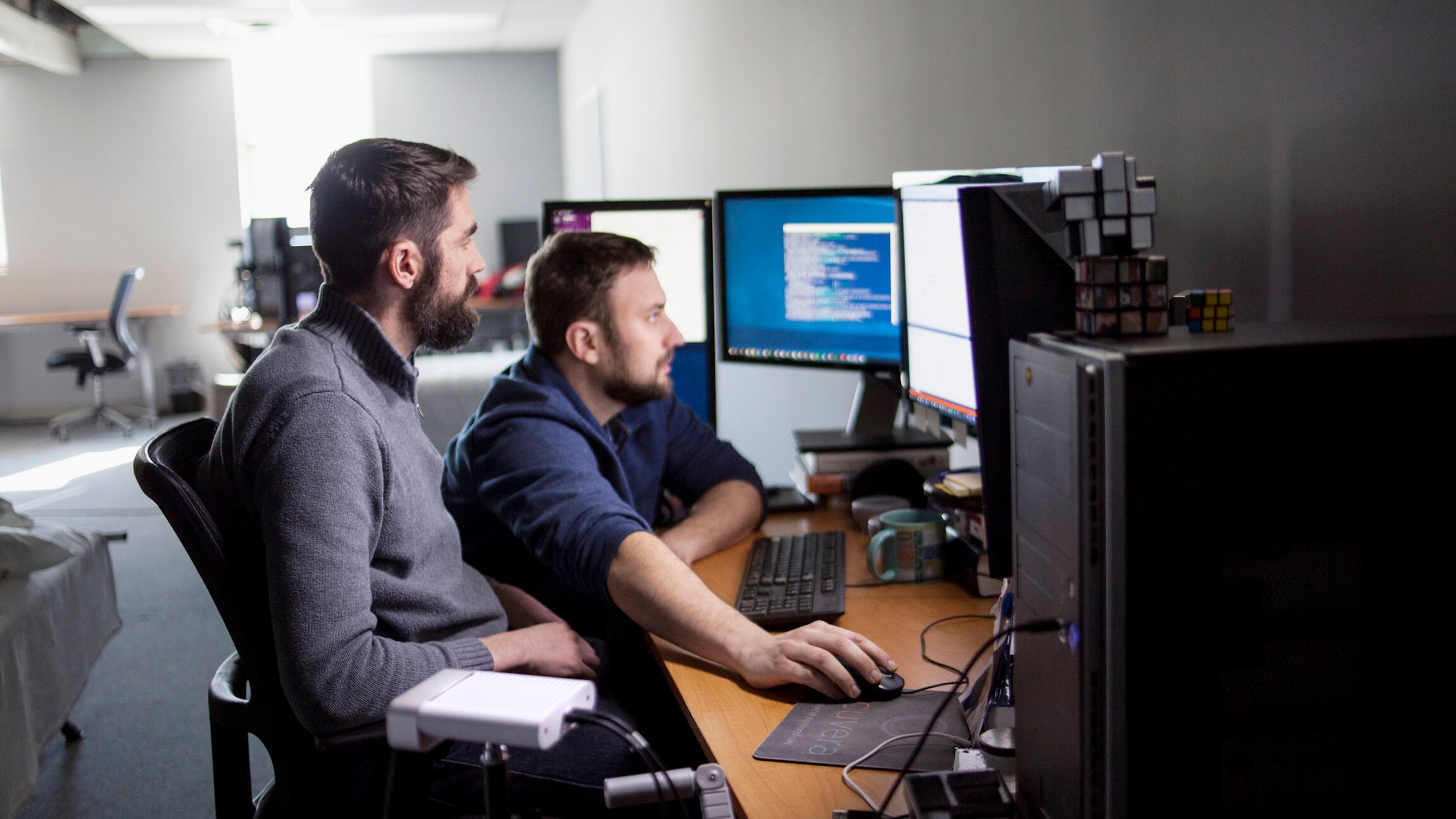 Two male engineers work in front of a multi-monitor setup in a darkened room.