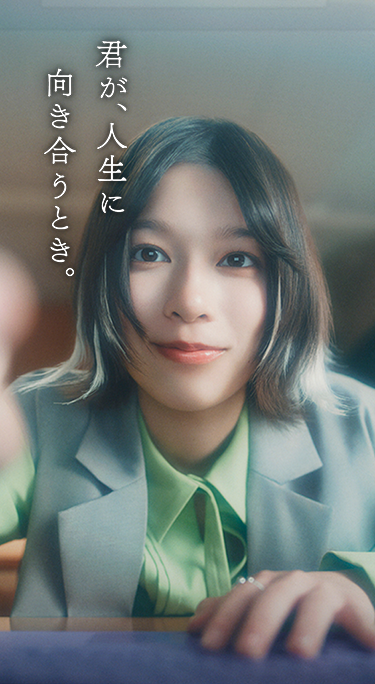 緑のシャツとグレーのベストを着た若い女性がカメラをじっと見つめている。手前は少しぼやけており、まるでデバイスを操作しているようだ。彼女の背後には、日本語の文字が反転表示されたノートパソコンの画面が見える。| 君が、人生に向き合うとき。