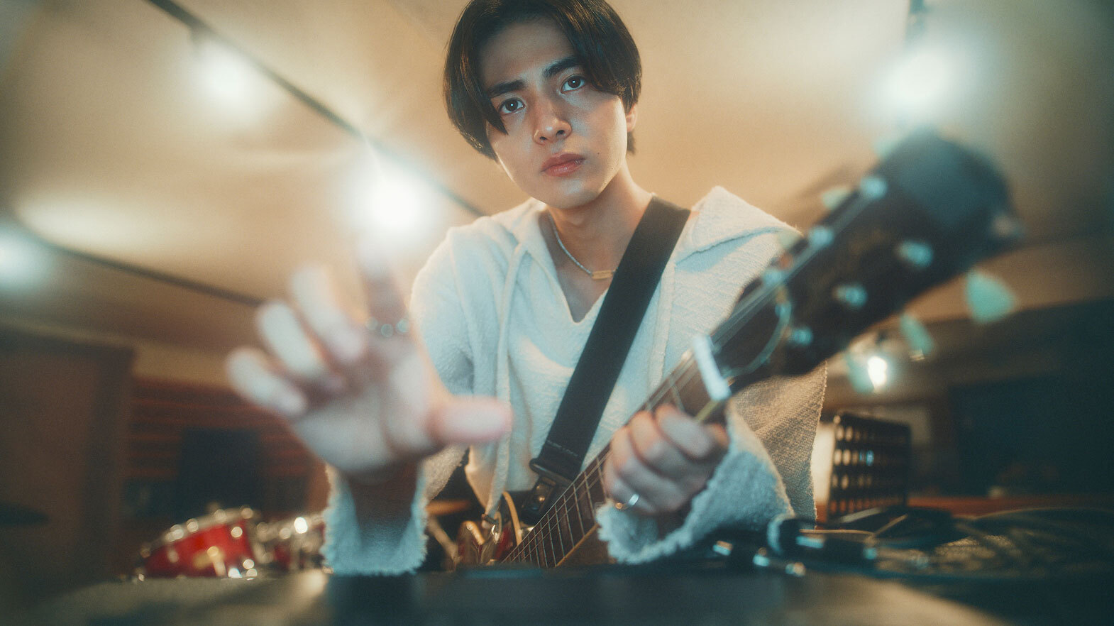 レコーディングスタジオでエレキギターを持ち、手前の機材に手を伸ばしている人物。
