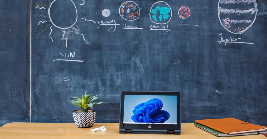 黒板の前のデスクに開いた状態のラップトップ PC やノート、植木鉢が置かれている様子