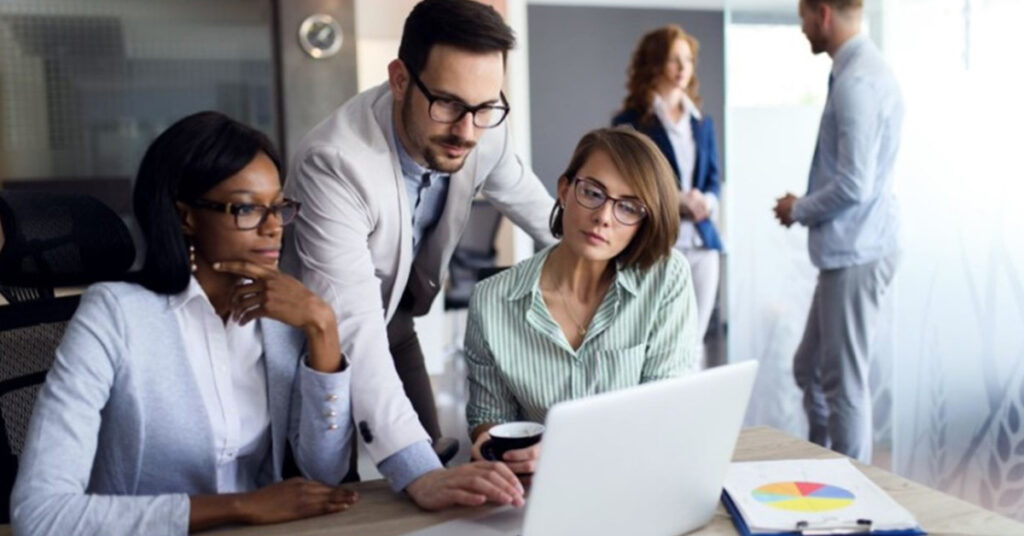 財務グラフが表示されたノート PC とタブレットをデスクで座って見ている 2 人の女性の財務専門家と、後ろから身を乗り出してノート PC 上で何かを操作している 1 人の男性の財務専門家。