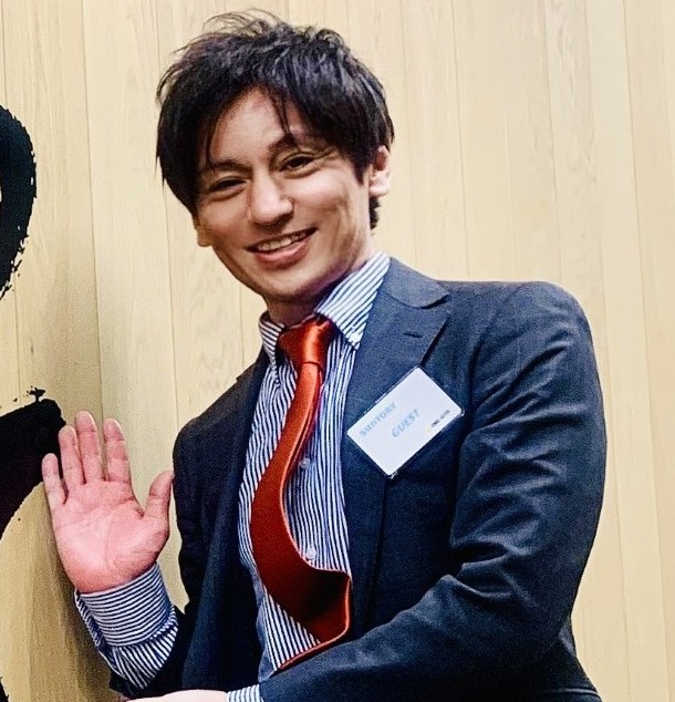 Person wearing a suit and tie, waving while standing indoors