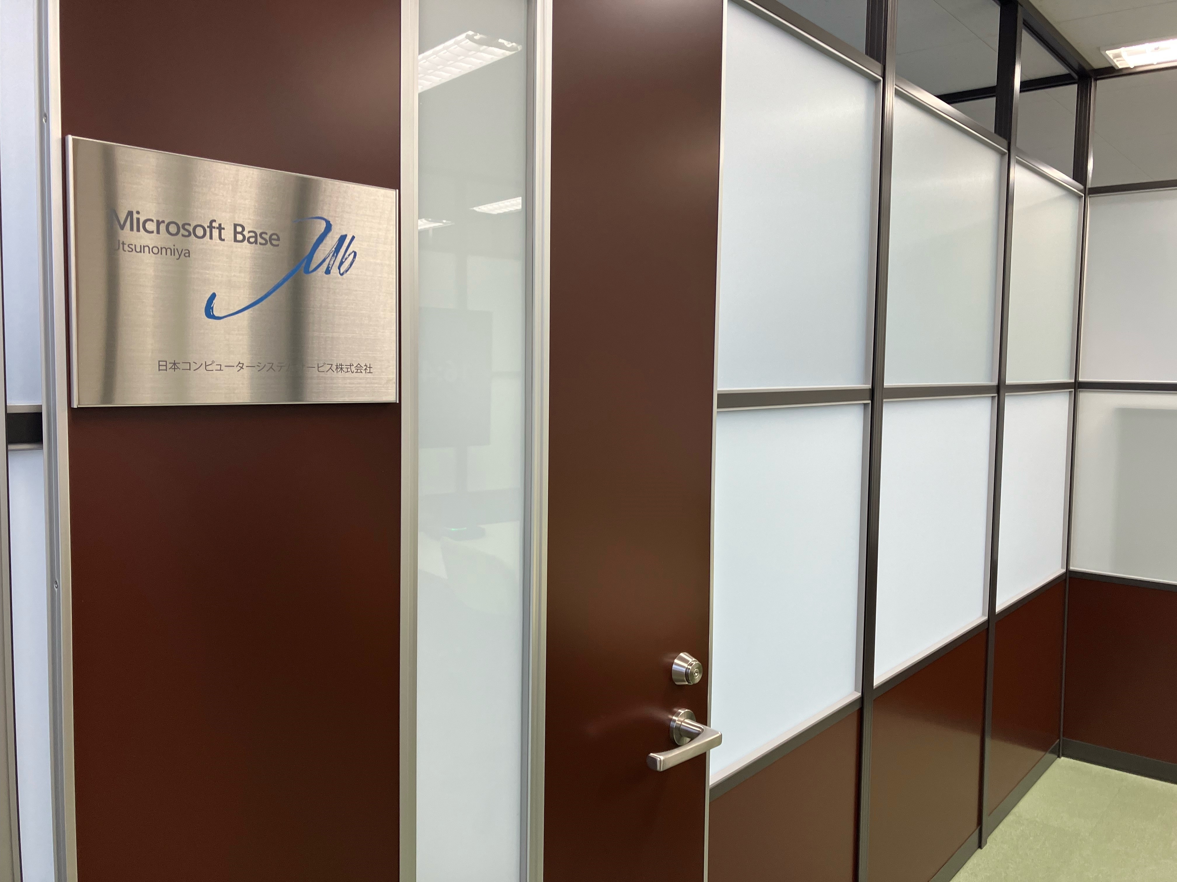 濃い茶色のドアと、「Microsoft Base Utsunomiya」と書かれたシルバーの看板があるオフィス入り口 | 日本コンピューターシステムサービス株式会社。