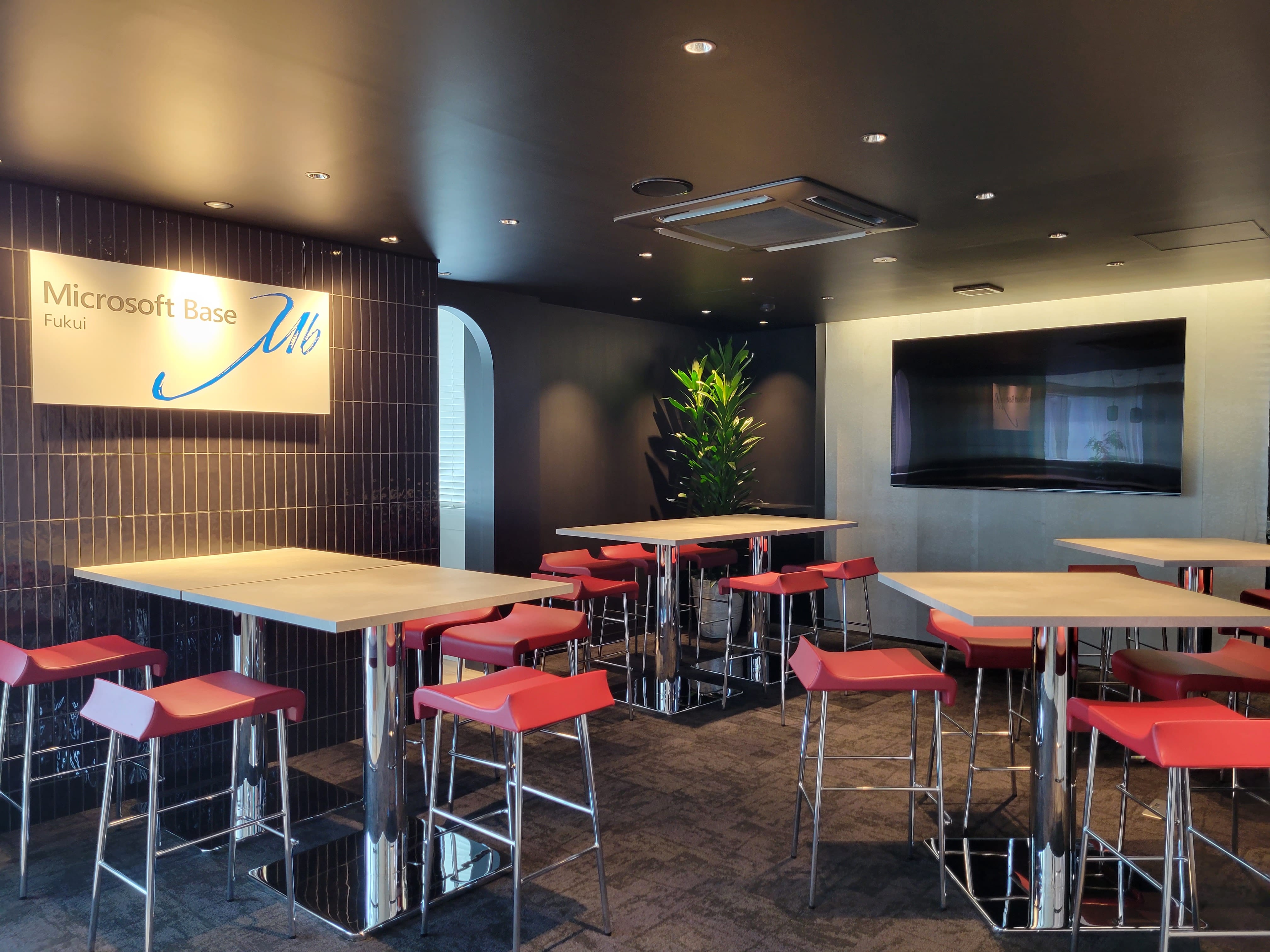 木製ハイトップテーブルと赤いバースツールが並ぶ席エリア。壁に「Microsoft Base Fukui」と表示。