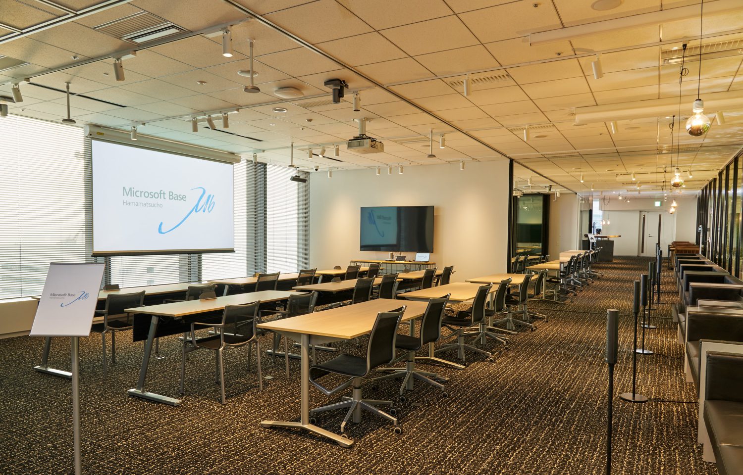 列になった机と回転椅子が前方のスクリーン（「Microsoft Base」表示）に向く研修室。側面にモニターとハイカウンター席あり | Hamamatsucho。