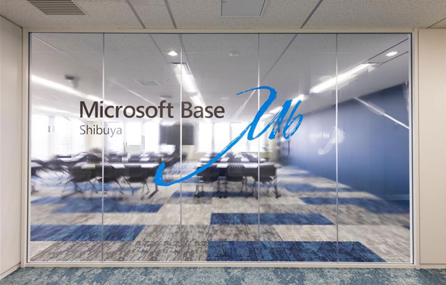「Microsoft Base Shibuya」と表示されたガラス壁越しに、列になった机とブルーのアクセントカーペットが見える研修室。
