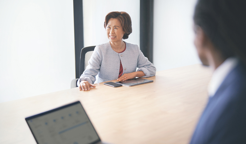 Uma mulher sentada a mesa com um celular e um tablet a frente. Do outro lado um homem com um notebook.