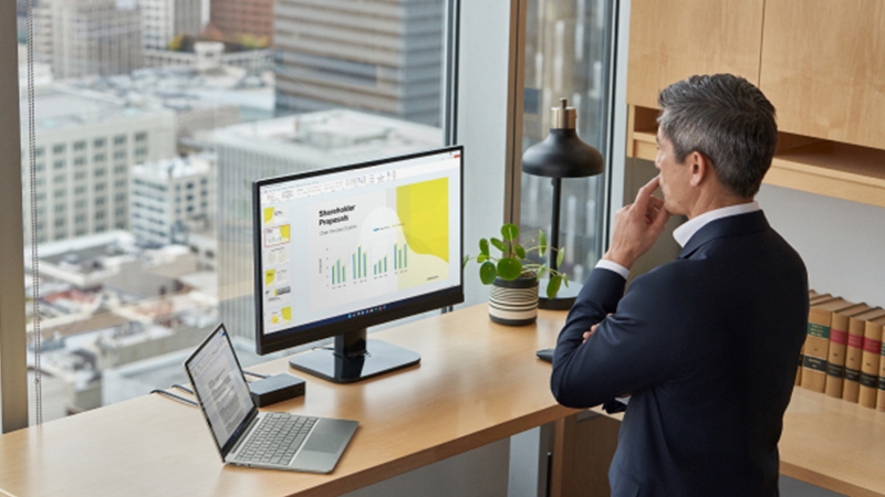 Um homem parado em frente a um monitor e um notebook em uma mesa com a cidade ao fundo pela janela.
